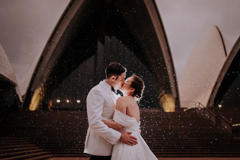 Bride and groom, Therese and Ash, sharing a romantic kiss in the rain on the steps of the Sydney Opera House during their wedding day.